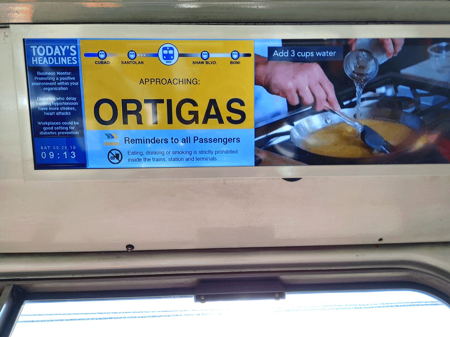 A monitor on mounted on the train's wall of MRT Line 3. The TUBE screen displays the next station, Ortigas, with an ad playing on its right and a news headlines column on its left, which also displays the time and date