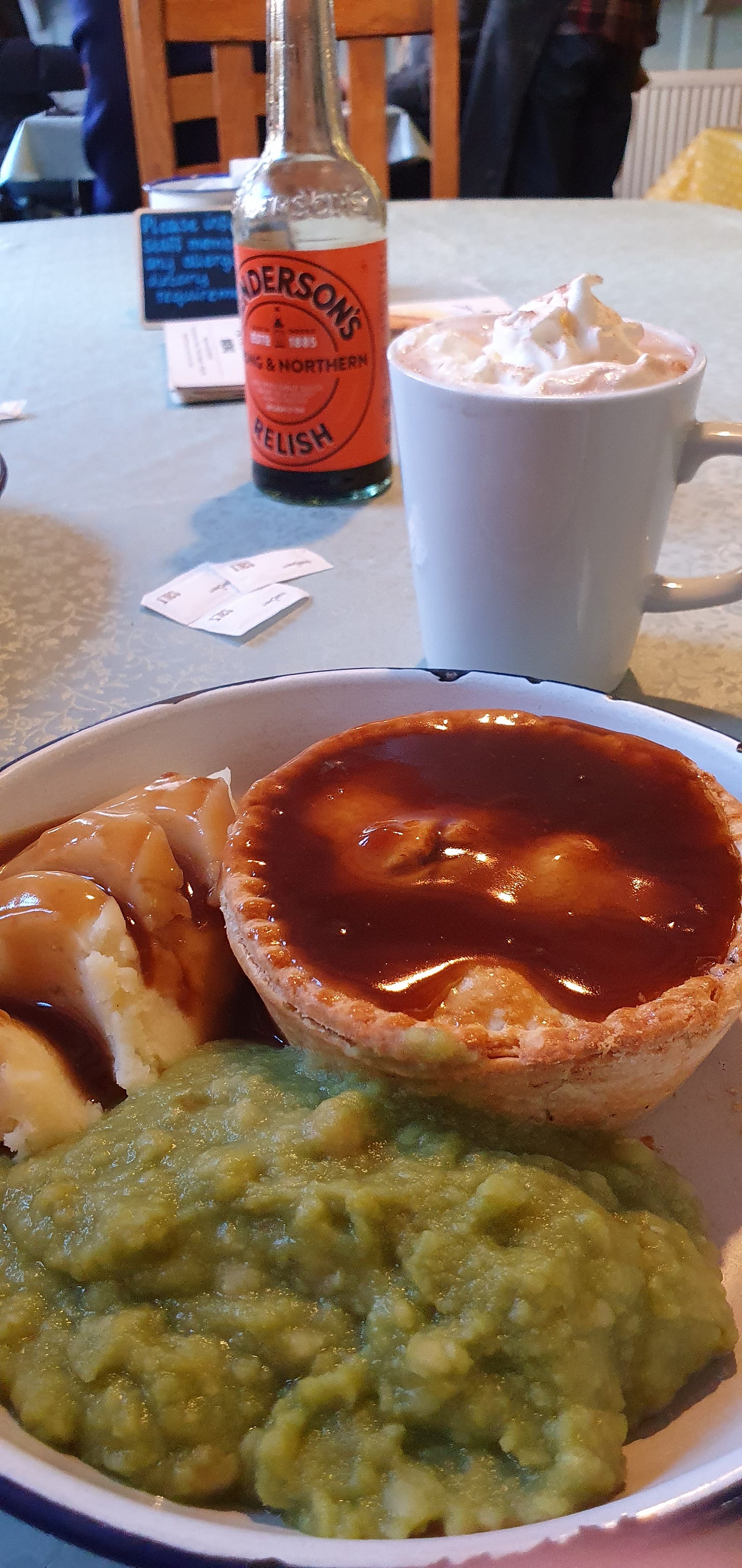 A portrait picture of an enamel bowl holding the discussed meat pie, pile of mashed potato, and portion of mushy peas. Behind it sits a mug of hot chocolate with whipped cream, and in the distance a bottle of Henderson's Relish can be seen.