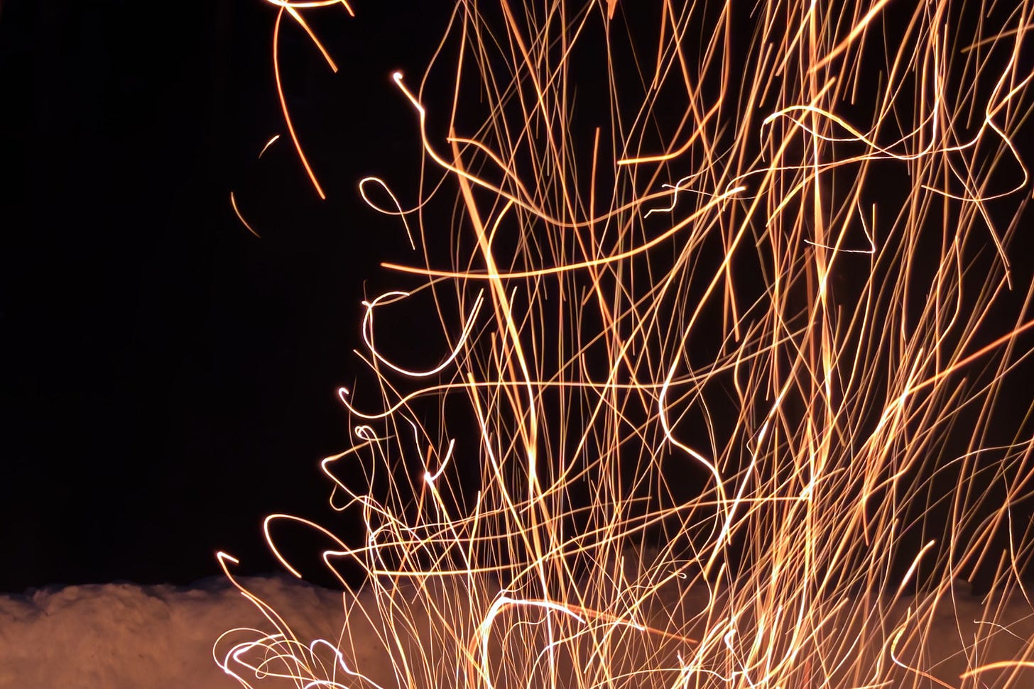 An image of a campfire at night is taken with a long exposure, so that the sparks flying up from the flames appear as a squiggly cluster of lines streaming orange against a black background. A low snowbank is visible behind. An image of a campfire at night is taken with a long exposure, so that the sparks flying up from the flames appear as a squiggly cluster of lines streaming orange against a black background. A low snowbank is visible behind.