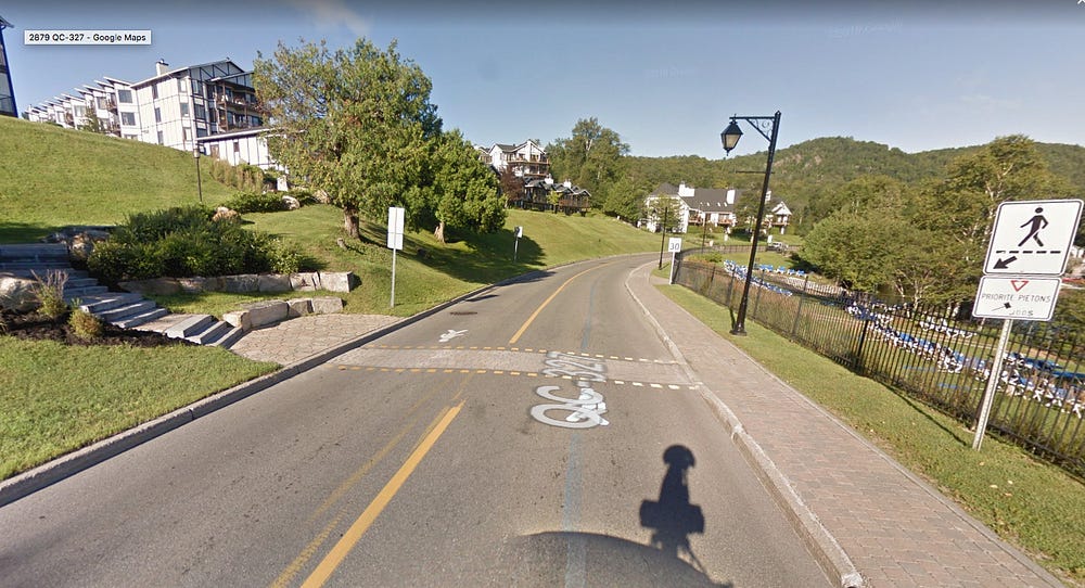 A two lane road with a sidewalk on one side and a marked brick crosswalk to access the business on the other side with a sign indicating pedestrian priority.