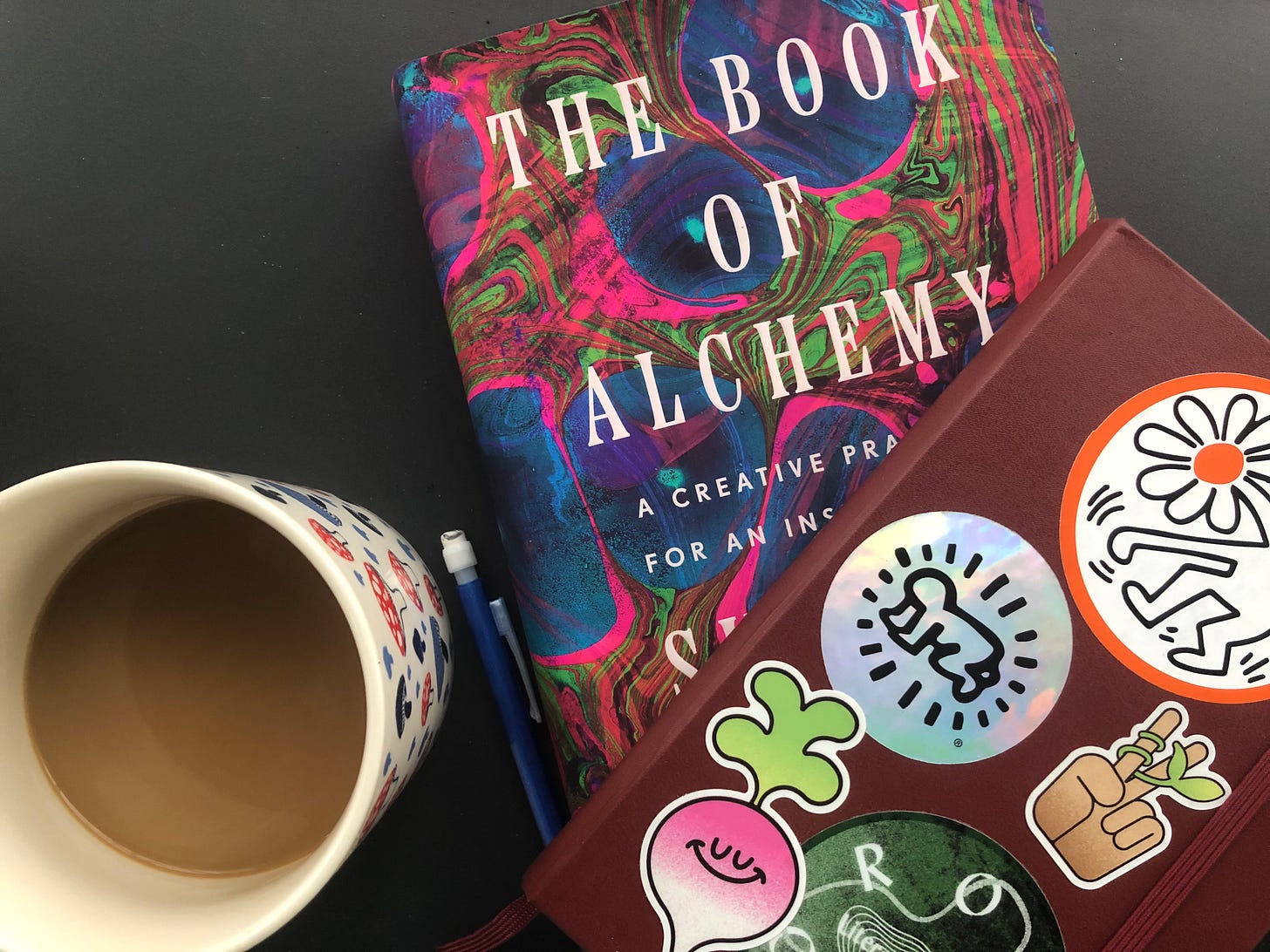 A mushroom-patterned mug half-full of coffee sits on a table next to a copy of Suleika Jaouad’s The Book of Alchemy and Emory’s sticker-covered notebook. A mushroom-patterned mug half-full of coffee sits on a table next to a copy of Suleika Jaouad’s The Book of Alchemy and Emory’s sticker-covered notebook.