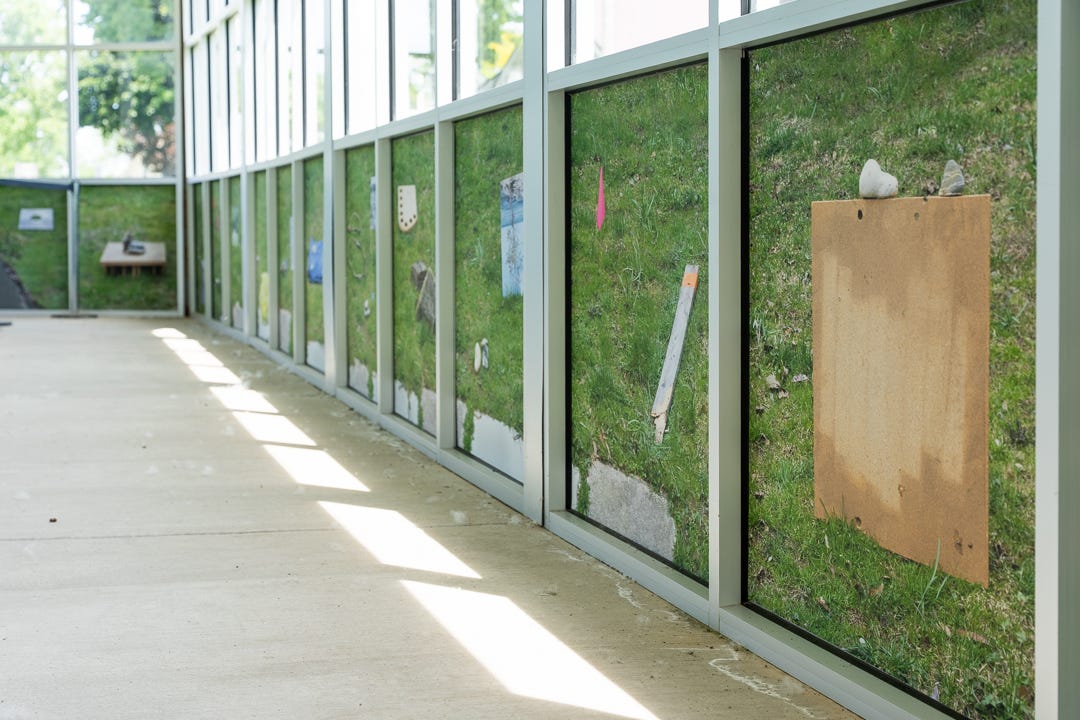 Large photos cover the glass windows of an ambiguous space. The photos show objects lying on the grass on the other side of the glass. Large photos cover the glass windows of an ambiguous space. The photos show objects lying on the grass on the other side of the glass.