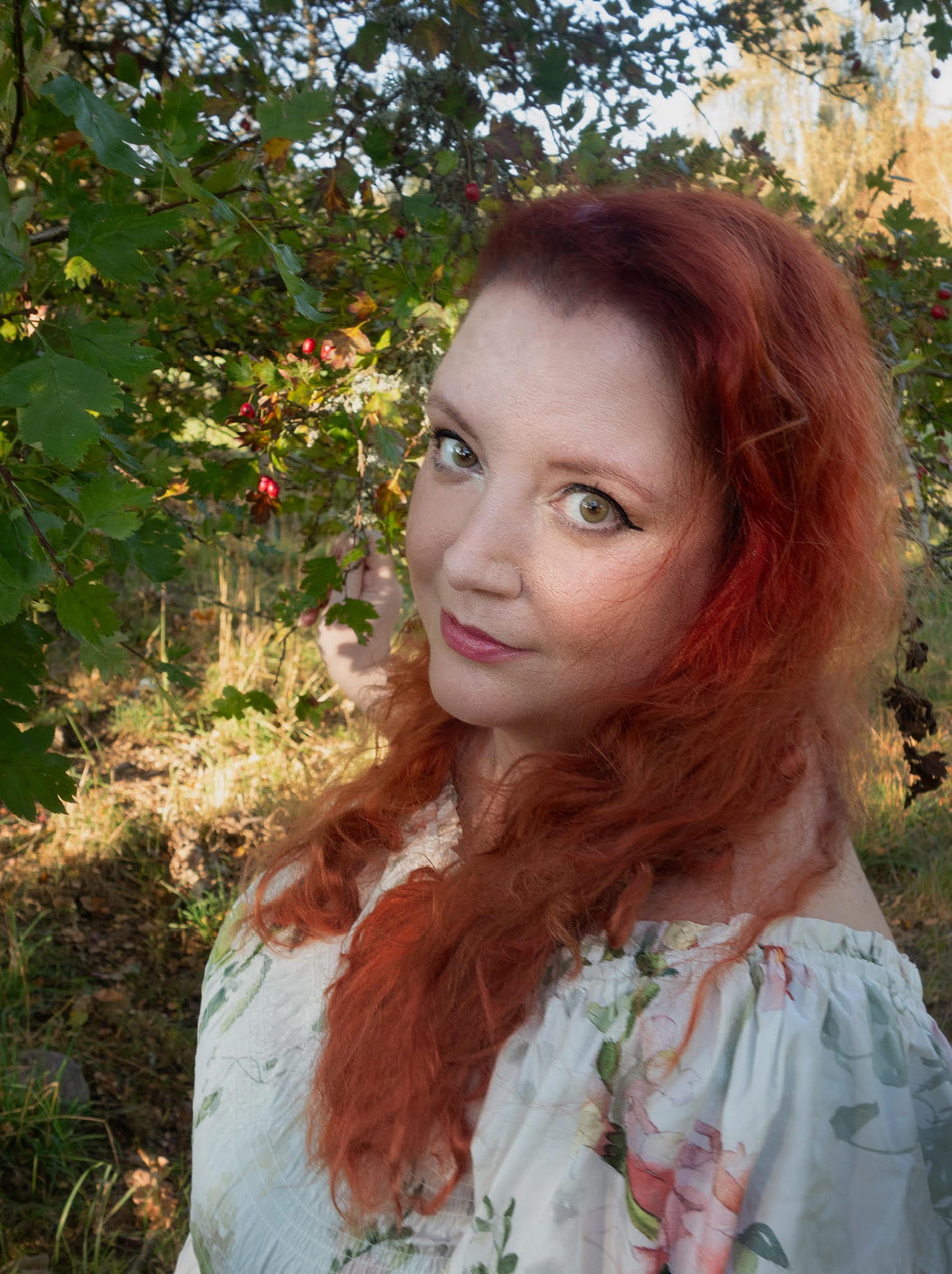 -Portrait of the author, Danai Christopoulou. They stand in front of a red berry tree, and wear an off-shoulder white dress with pink and green blooms. It's summer, and the sunset rays fall on their long wavy red hair.