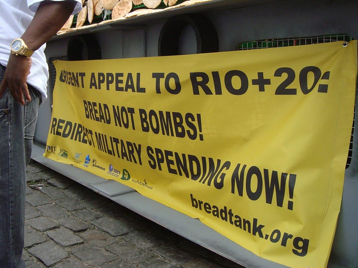 First image: A close-up of a bright yellow banner attached to the side of a grey military vehicle. The black words in bold capital letters read: "URGENT APPEAL TO RIO+20 BREAD NOT BOMBS! REDIRECT MILITARY SPENDING NOW!" The banner is also adorned with supporters' logos and with the web address "breadtank.org"; second image: a man is preparing round flatbreads at a table with a photographer next in line, behind him a man with a microphone is speaking to a crowd of people out of view, with people beside him; in the background is a military tank adorned with round flatbreads and the yellow BREAD NOT BOMBS banner.