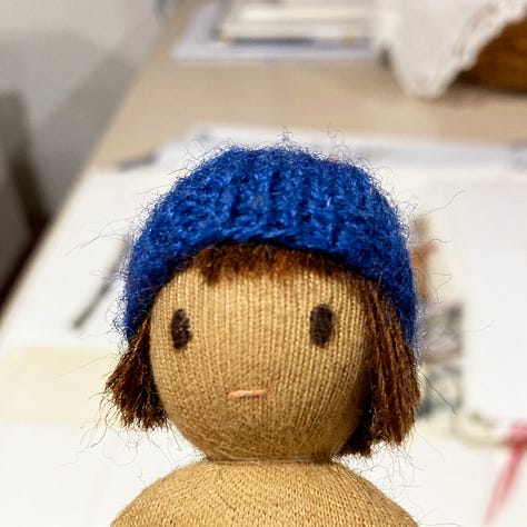 Three photos showing before, during and after the process of sewing brown wool hair onto a small handmade cloth doll made by Illustrator HannahSangjuinetti. The doll has coffee coloured skin and a blue knitted hat and is shown alongside the sketch of an illustration she will eventually be part of.