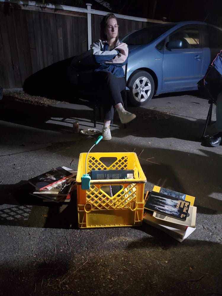 A book light illuminates a yellow milk crate filled with books and surrounded by stacks of books. It is night-time in a driveway. A person in glasses sits beyond the crate in a folding camp chair. A book light illuminates a yellow milk crate filled with books and surrounded by stacks of books. It is night-time in a driveway. A person in glasses sits beyond the crate in a folding camp chair.