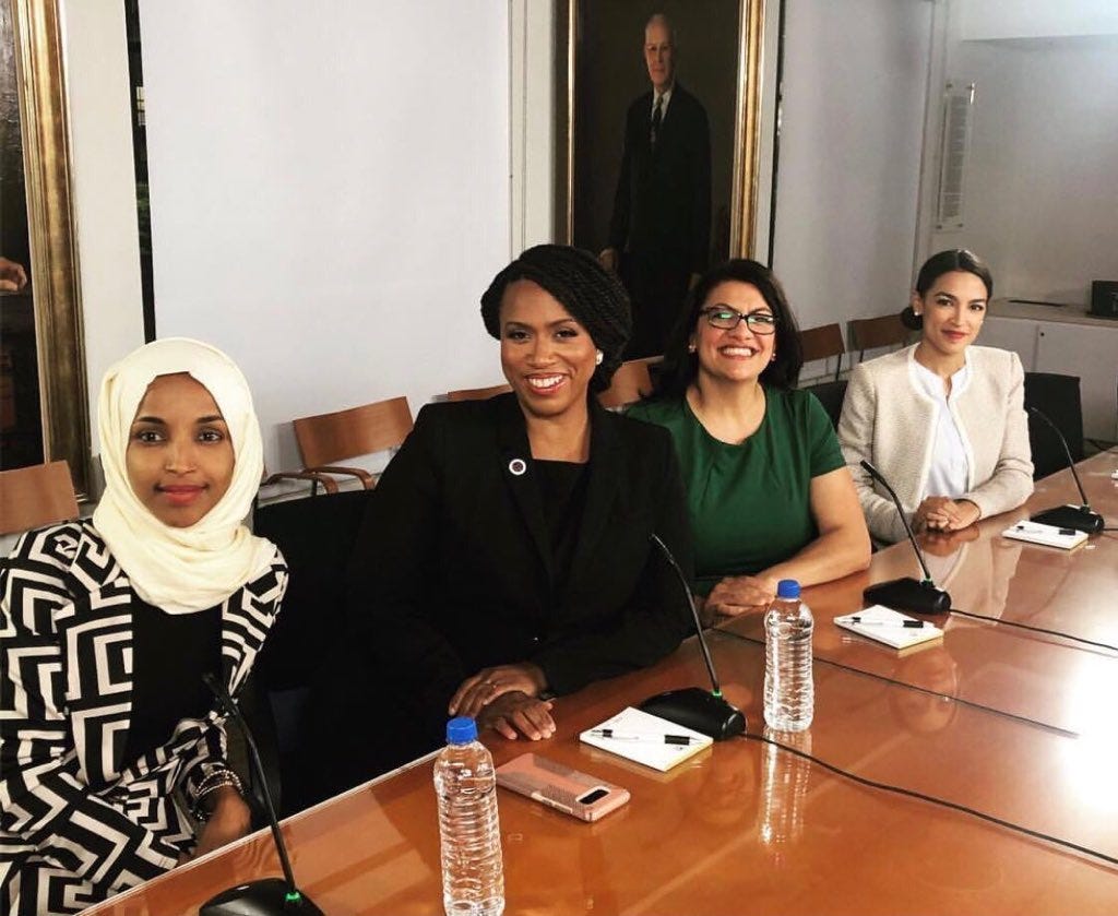 Rep. Ilhan Omar, Rep. Ayanna Pressley, Rep. Rashida Tlaib, Rep. AOC sitting next to each other leaving on a table