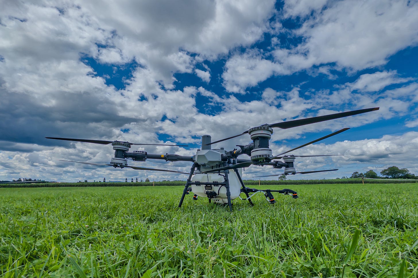Auf einer Wiese steht eine grosse landwirtschaftliche Mehrrotor-Drohne für Pflanzenschutz oder Ausbringung. Die DJI Agras T100 hat vier koaxiale Doppelrotoren, ein zentrales Gehäuse mit Tank und ein robustes Landegestell. Durch die tiefe Perspektive wirkt die Drohne technisch und massiv; darüber spannt sich ein Himmel mit grossen weissen und dunkleren Wolken.