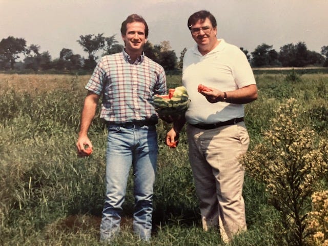 Two men standing in a field holding fruits

AI-generated content may be incorrect.