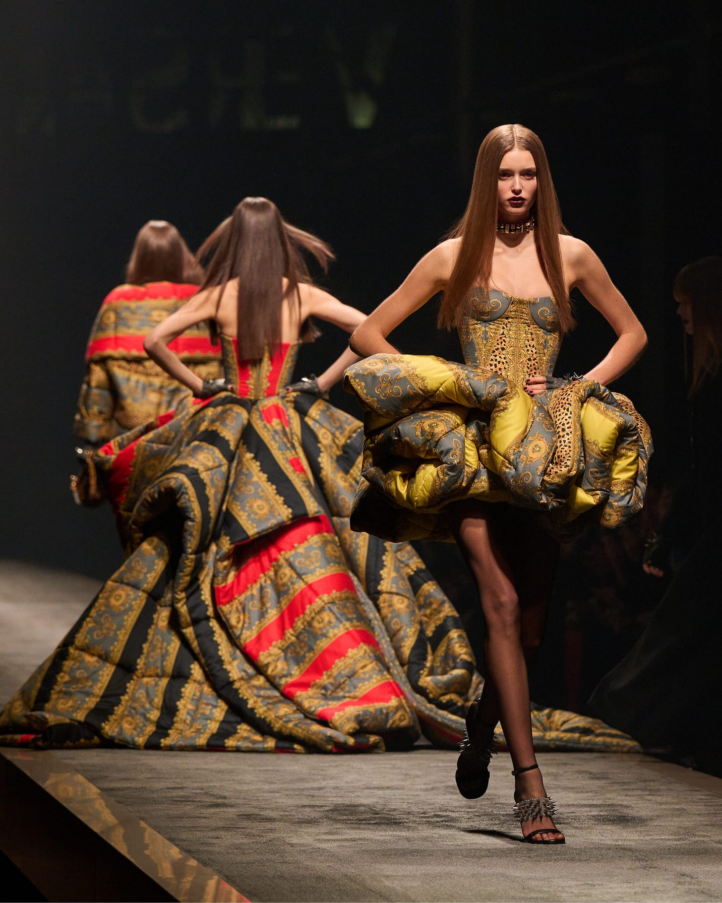A model wearing a densley printed mini dress walks the runways as two other models in similarly printed gowns walk in the opposite direction behind her.
