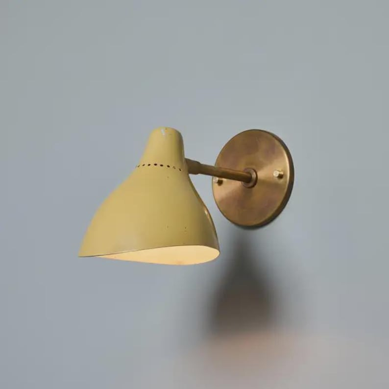 May include: A wall-mounted light fixture with a pale yellow conical shade and a brass-colored circular base. The shade has a series of small holes near the top. The fixture is mounted on a light blue wall. May include: A wall-mounted light fixture with a pale yellow conical shade and a brass-colored circular base. The shade has a series of small holes near the top. The fixture is mounted on a light blue wall.