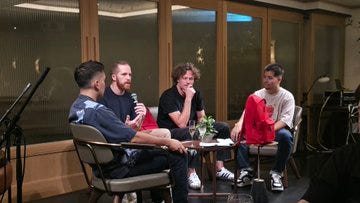 Four individuals sitting in chairs, engaged in conversation around a small table with a plant. A microphone and camera equipment are visible, capturing the discussion. A red backpack is placed on one chair. Large windows with wooden frames and a modern indoor setting with tiled walls are in the background. Other people are seated at tables, eating and talking in a spacious room with wooden accents and large windows.