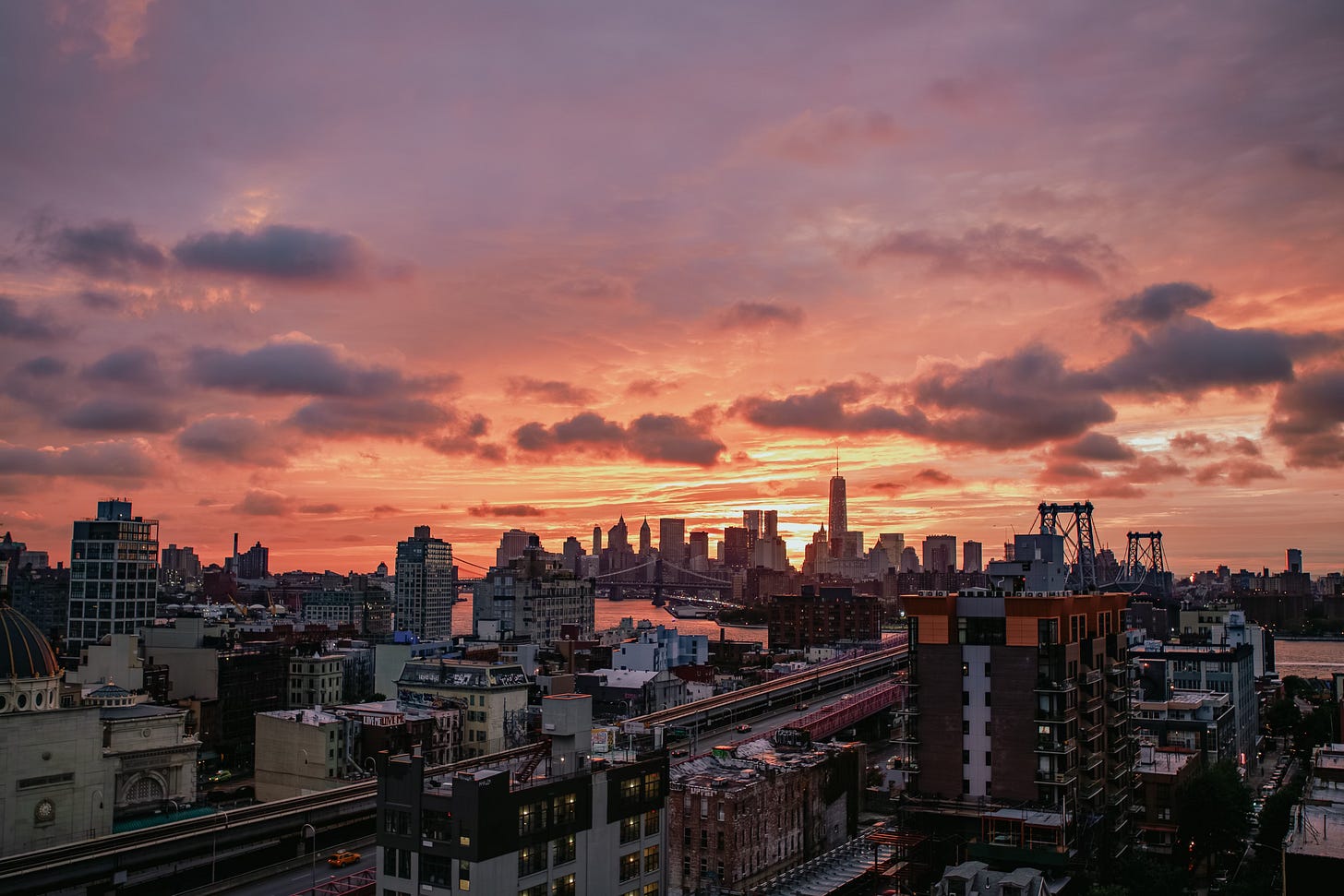 A wide, elevated view of the Lower Manhattan skyline under a dramatic, cloudy sunset. The clouds are colored orange and pink. In the foreground are Brooklyn rooftops and an elevated train line, with the Williamsburg Bridge visible on the right.