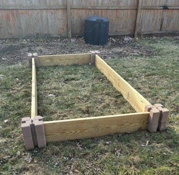 An 8' by 4' garden bed sits empty in a yard, with a weathered wooden privacy fence in the background.