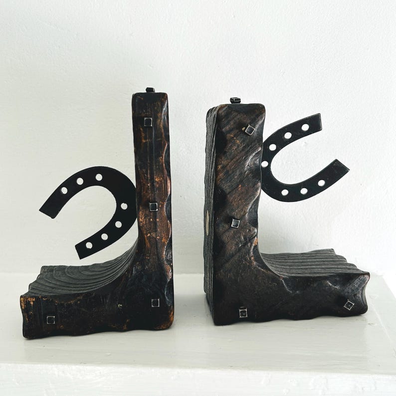 May include: A pair of rustic wooden bookends with metal horseshoe accents. The bookends are dark brown and have a rough, textured surface. The horseshoes are black and have a simple design.