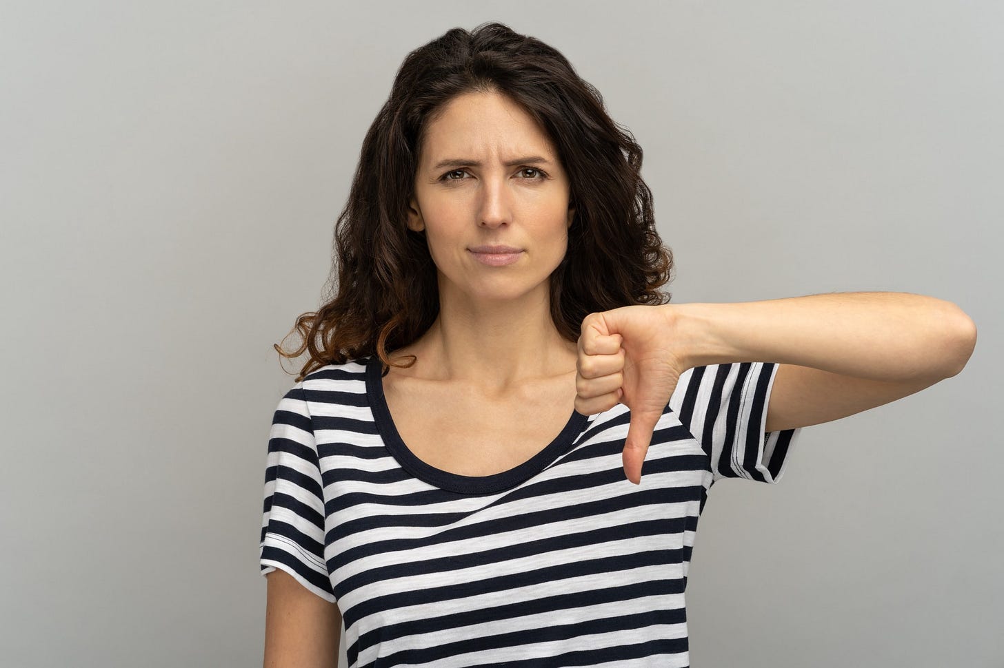 A woman with long dark hair scowls as she gives a thumbs down gesture.