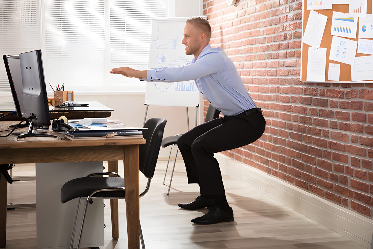 Man dressed in work attire doing a standing squat in front of his desk. Man dressed in work attire doing a standing squat in front of his desk.