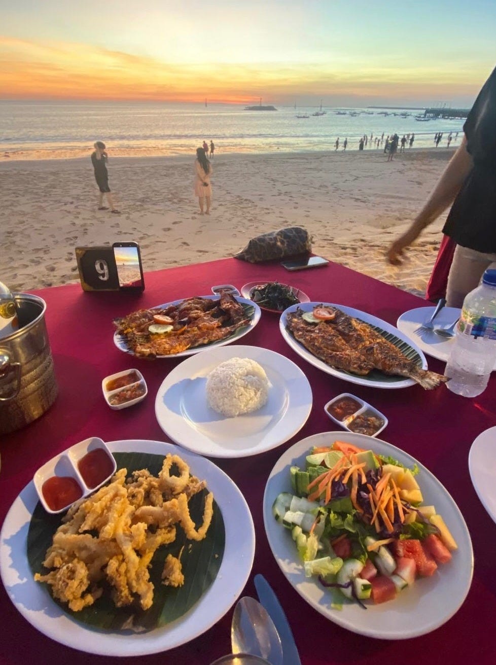 Seafood dinner set on a beach table at sunset in Bali, with multiple dishes and the ocean in the background.