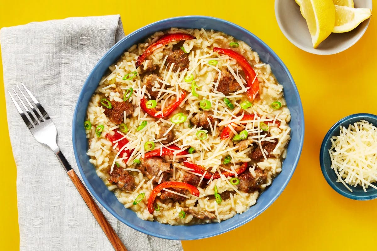 A bowl of pork sausage pepper risotto, next to a napkin, fork, grated cheese, and lemon wedges