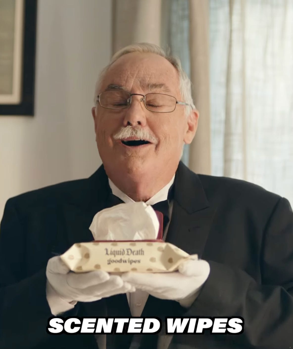 Older man in a tuxedo and white gloves holding a box of Liquid Death-branded scented wipes, with his eyes closed as if smelling them. Large text at the bottom reads, “SCENTED WIPES.”