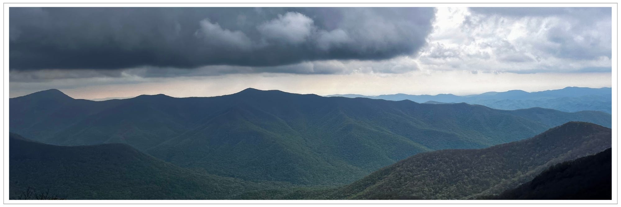 dense, dark cloud hovering over mountain ridges, dusky light in distance, newly greening trees in the valleys of rolling mountain ranges