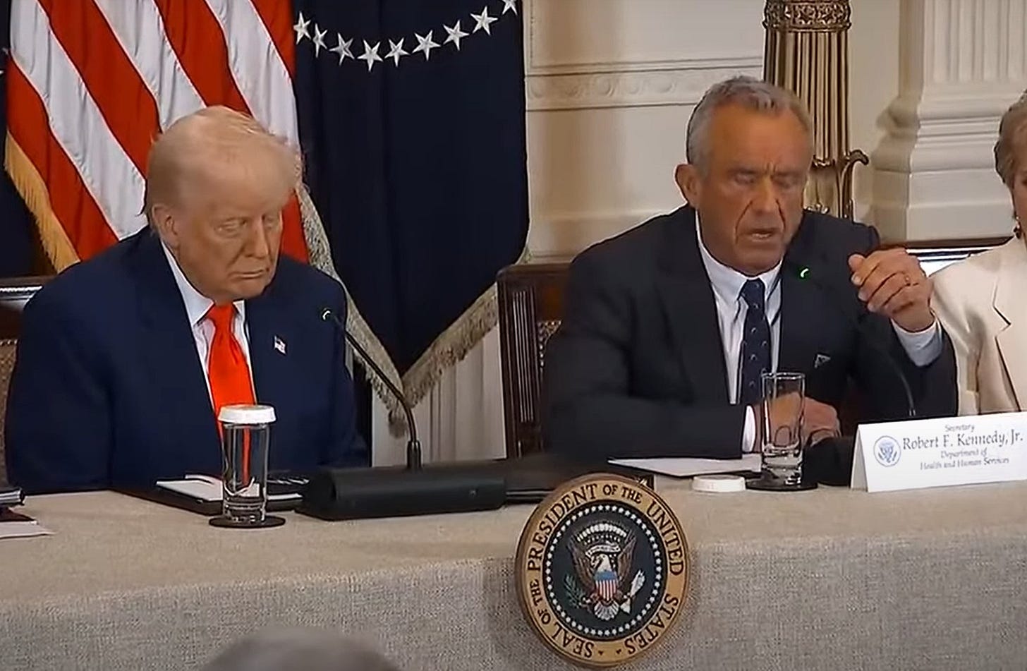 Video screenshot of Donald Trump and RFK Jr sitting at a dais during a White House event. The US presidential seal is affixed to the table in front of Trump. 