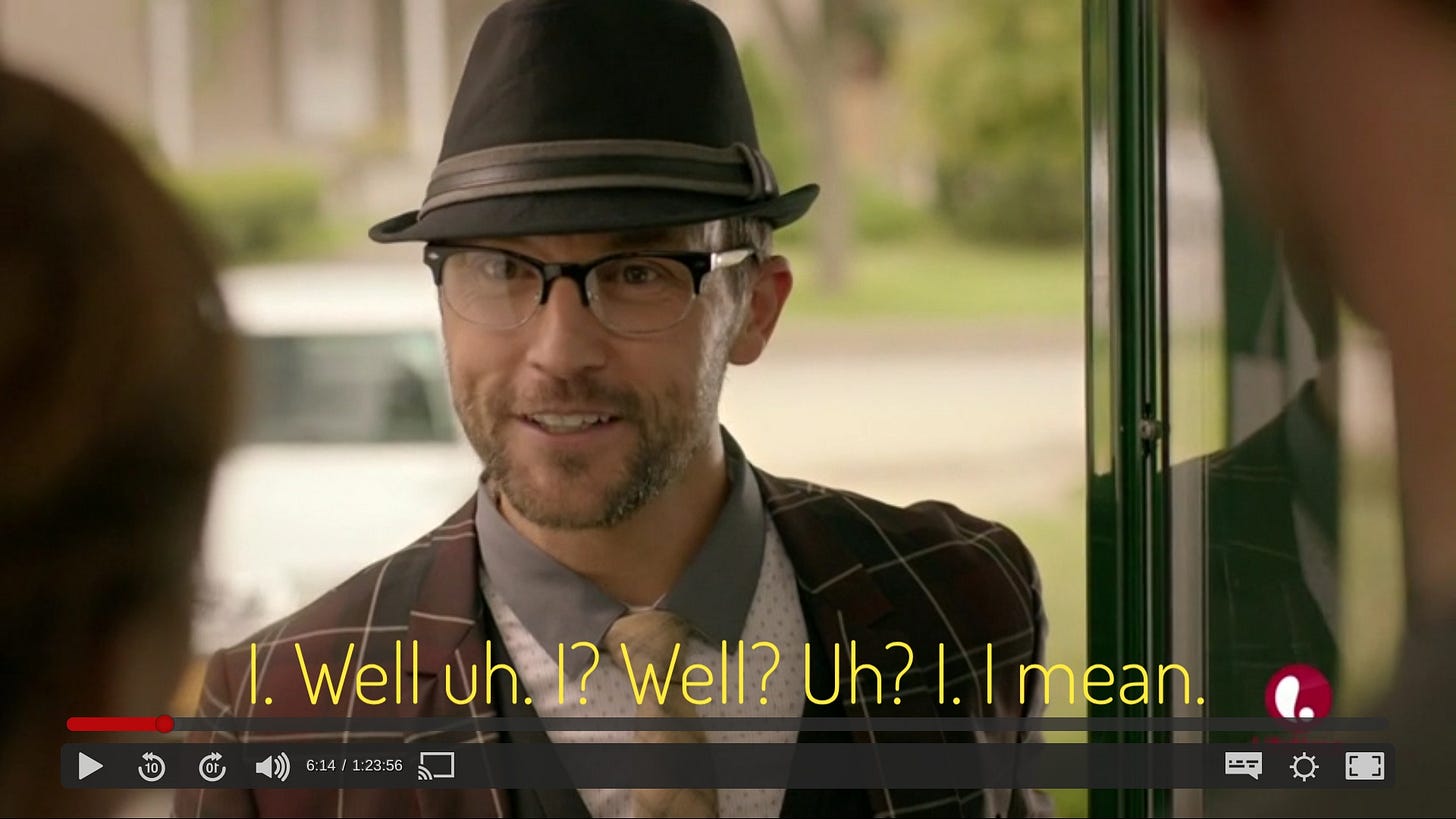 Simon, a white man with stubble, smiling and wearing a trilby, plaid jacket, shirt with little dots and blue collar, and yellow skinny tie. Captioned "I. Well uh. I? Well? Uh? I. I mean."