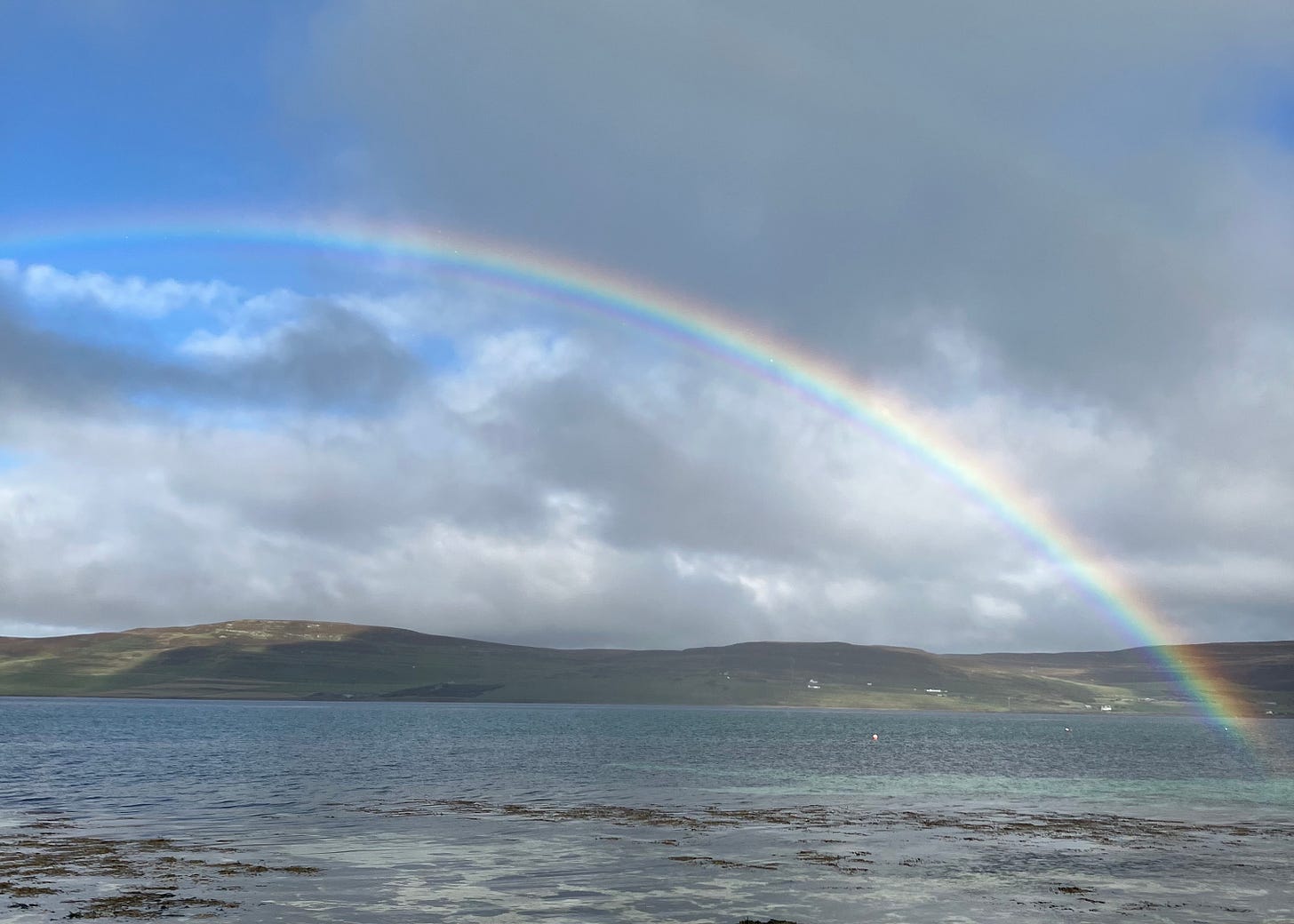 A photo of a placid sea with a dark green island in the distance. The sky is blue, but dark clouds are shifting over the landscape, darkening it in parts. A rainbow arches over the scene