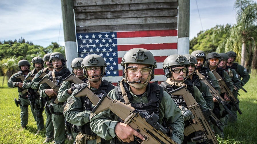 A group of ICE officers in military-style uniforms poses on a grassy training field with their guns, helmets, and body armor, in front of a climbing obstacle painted with a US flag. They all have very serious tough-guy expressions on their faces. A group of ICE officers in military-style uniforms poses on a grassy training field with their guns, helmets, and body armor, in front of a climbing obstacle painted with a US flag. They all have very serious tough-guy expressions on their faces.