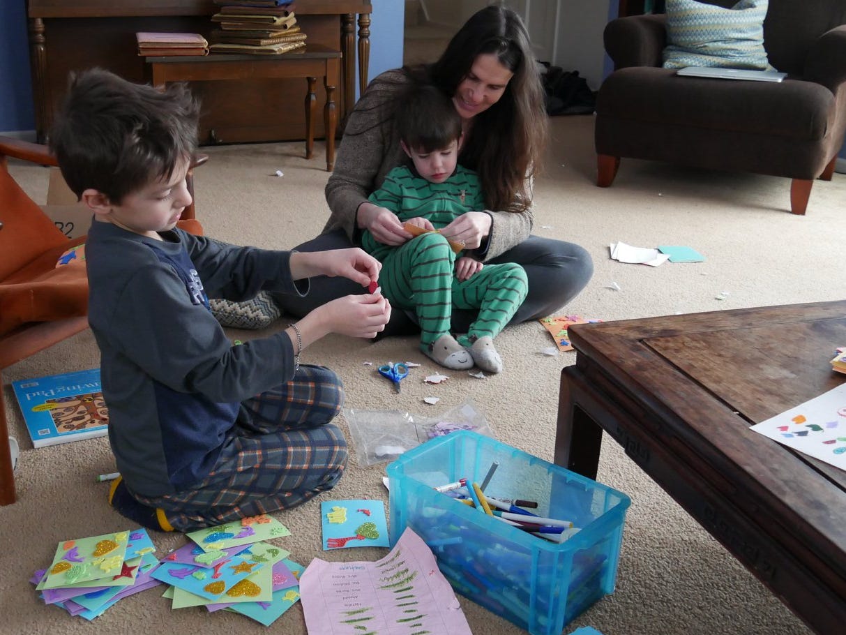 A boy sits on the carpet with piles of cards full of spongy stickers shaped like stars, hearts, and butterflies. There is a list of names, crossed out by his name and a box of markers. Behind him a woman has a toddler on her lap. He too is working on stickers on cards. It is daytime. They are in a living room. A boy sits on the carpet with piles of cards full of spongy stickers shaped like stars, hearts, and butterflies. There is a list of names, crossed out by his name and a box of markers. Behind him a woman has a toddler on her lap. He too is working on stickers on cards. It is daytime. They are in a living room.
