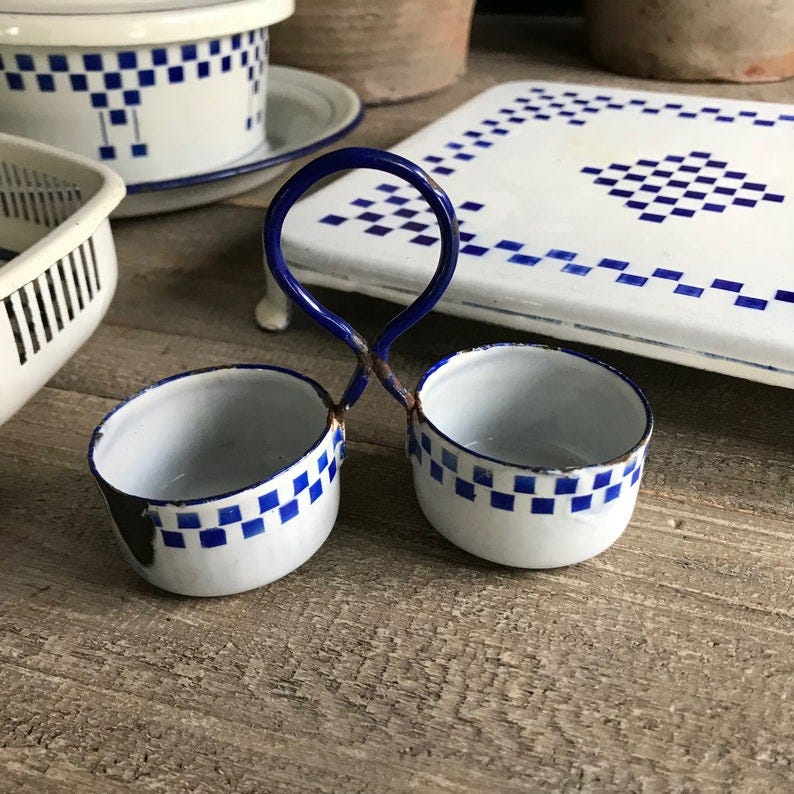 May include: Two small white enamelware bowls with blue checkered trim attached to a metal handle. The bowls are empty.