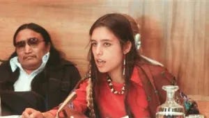 Winona as an 18 year old, leaning forward as she talks at a table. She has long brown braids coming down behind her ears. She is wearing a red shirt that looks like traditional native clothing, with a bead necklace. There is a native man sitting behind her watching her as she speaks. She is intently speaking and looking at the person in front of her, off camera.