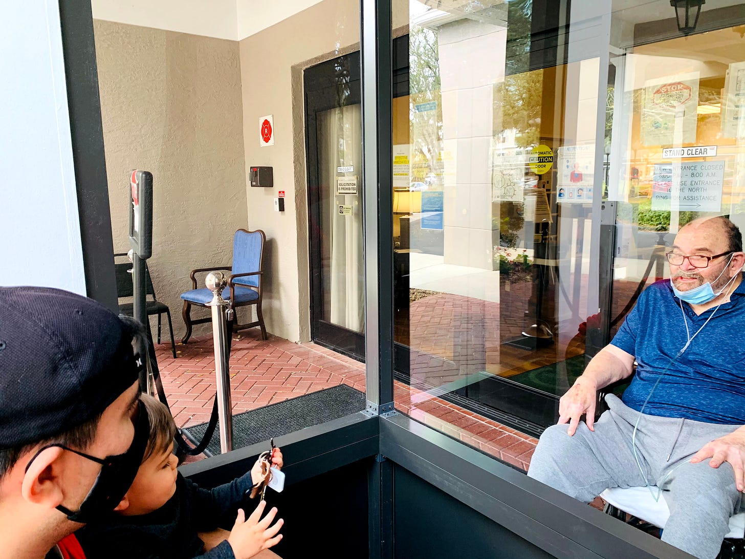 An old man with a face mask and oxygen tubes sits and looks at a dad with a baby from behind glass.