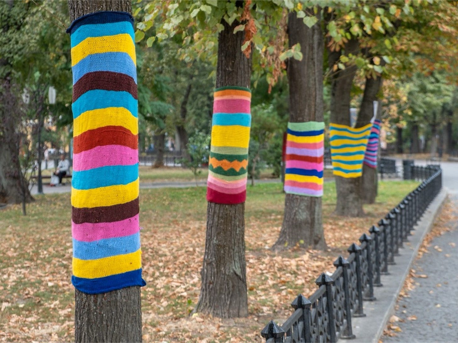 Trees wrapped in colorful knit patterns in a park. Autumn leaves on the ground, black wrought iron fence lining a pathway. Vibrant mood.