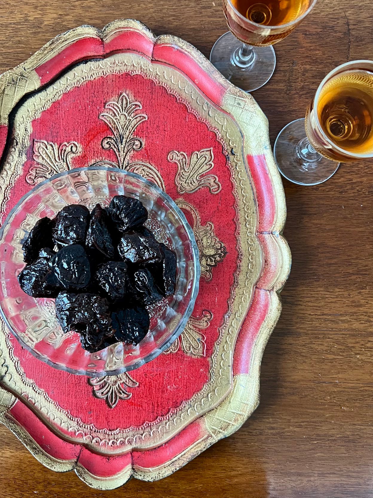 Overhead shot of dish of dried prunes on a red tray with gold decorations & 2 glasses of sherry