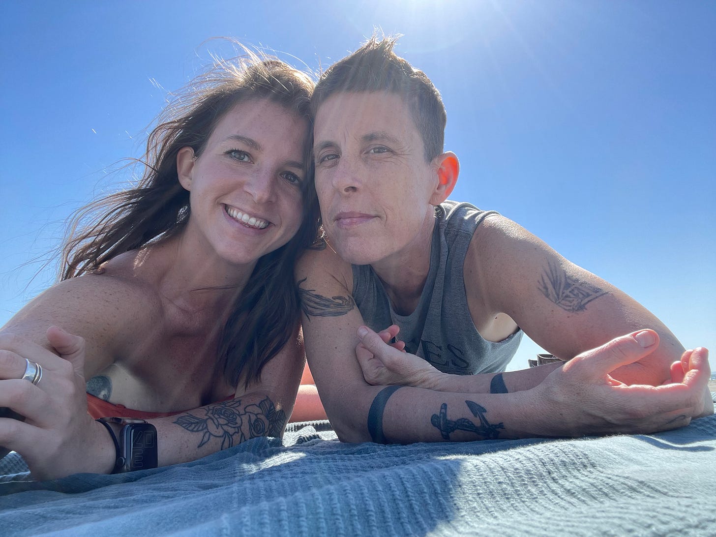 Meg and Andrea, laying on a towel at the beach, heads pressed together, smiling into the camera, joyful, in love, and alive. Meg and Andrea, laying on a towel at the beach, heads pressed together, smiling into the camera, joyful, in love, and alive.