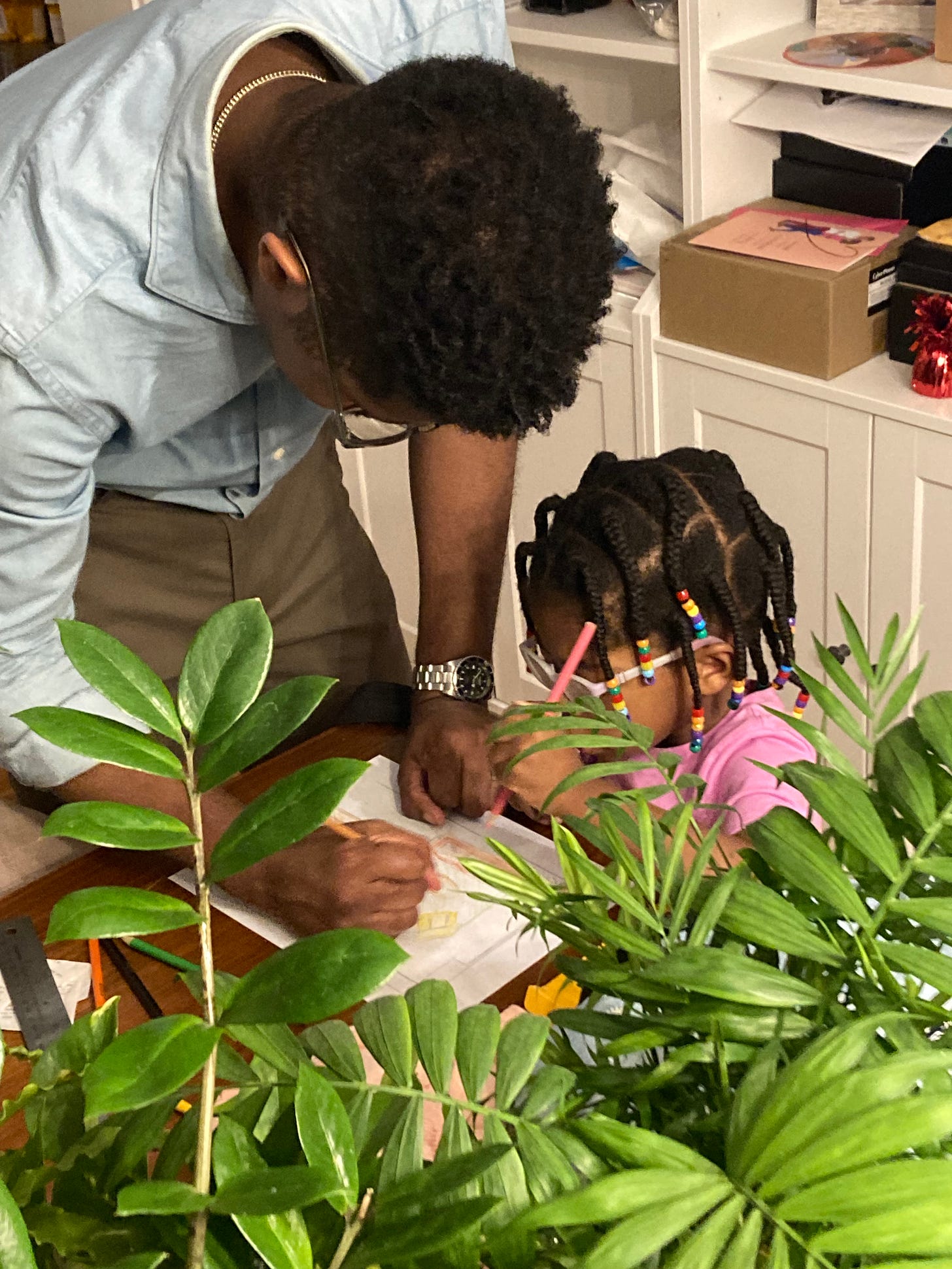 Picture of a Black dad and a Black toddler girl drawing. Houseplants are in the foreground.