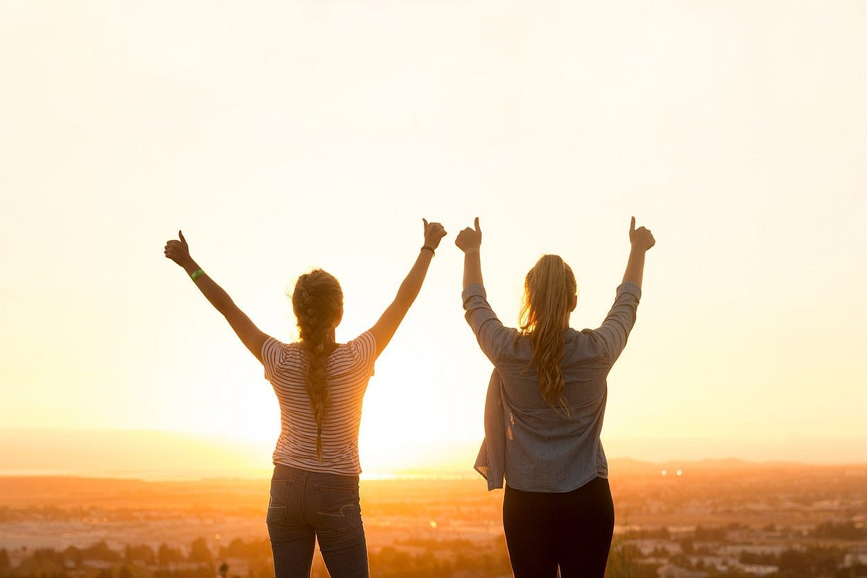 Image of two girls seen from the back, their arms in the air, thumbs up and in the background you see the sunset.