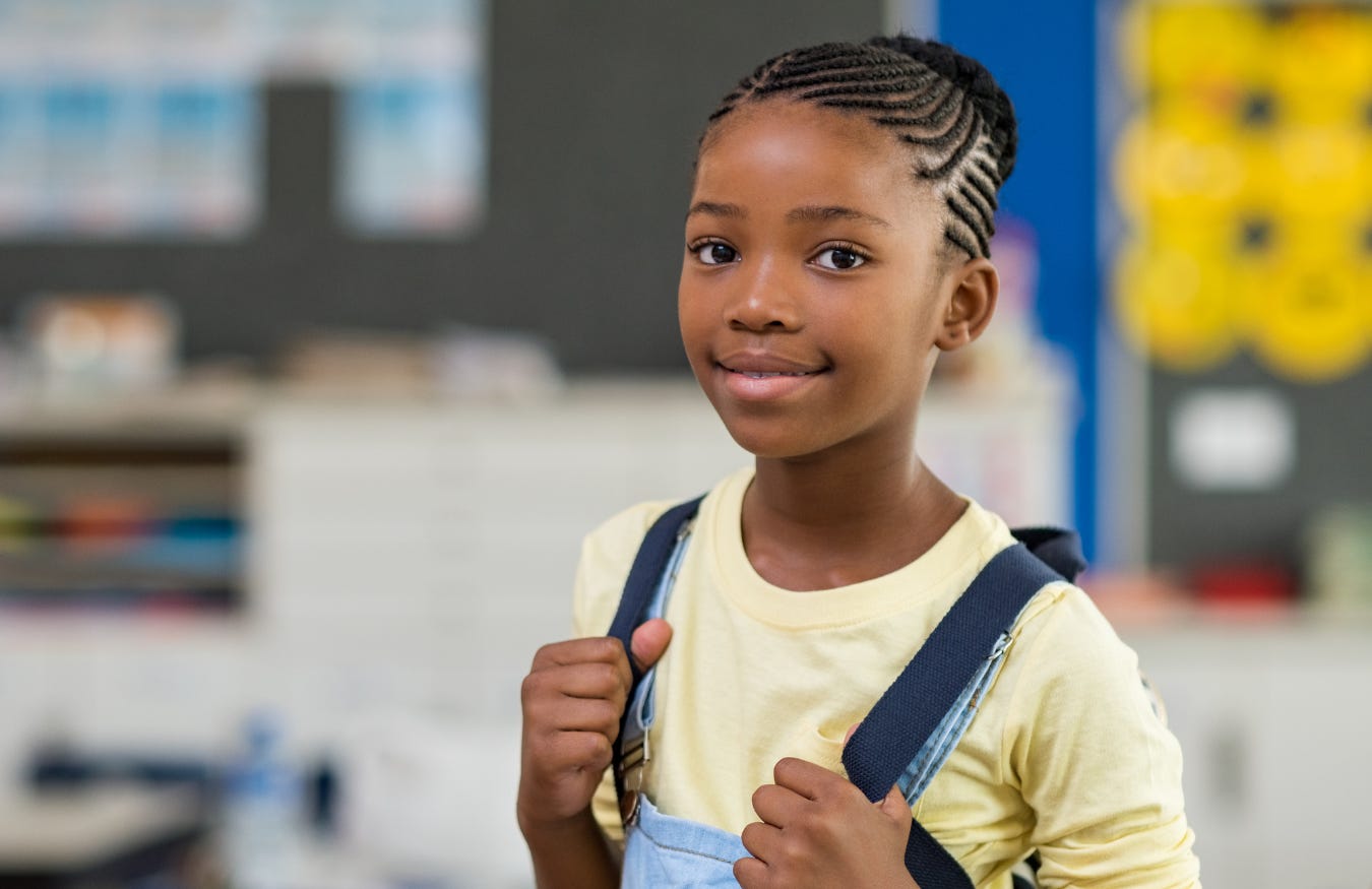 10-12 year old girl smiling gently at camera while standing in a classroom holding her backpack.