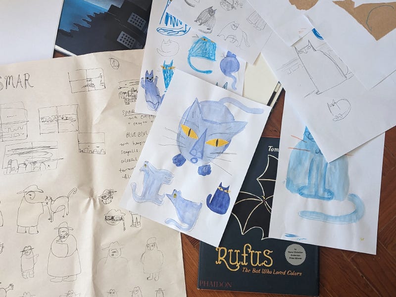Photo of scattered papers and sketches of blue cats on the floor, showing the process of picture book making.
