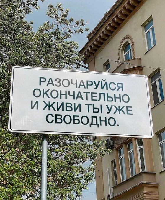 White rectangular signboard mounted on a metal pole in front of a two-story beige building with arched windows and a sloped roof under a partly cloudy sky. Green leafy trees frame the left side. The signboard text in bold black uppercase Russian reads РАЗОРЯЙ ЭКОНОМИЮ И ТЫ РАЗРУШИШЬ ВОЙНУ, translating to Destroy the economy and you will destroy the war. Additional buildings and trees are visible in the background.