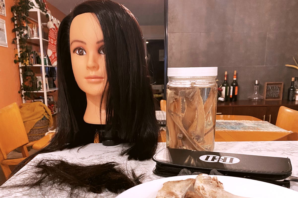 Table scene with a doll head mannequin (Brenda) with long dark hair, next to a clear glass jar filled with preserved animal bones, and a small pile of dark hair. Shows unique and eclectic home decor. Table scene with a doll head mannequin (Brenda) with long dark hair, next to a clear glass jar filled with preserved animal bones, and a small pile of dark hair. Shows unique and eclectic home decor.