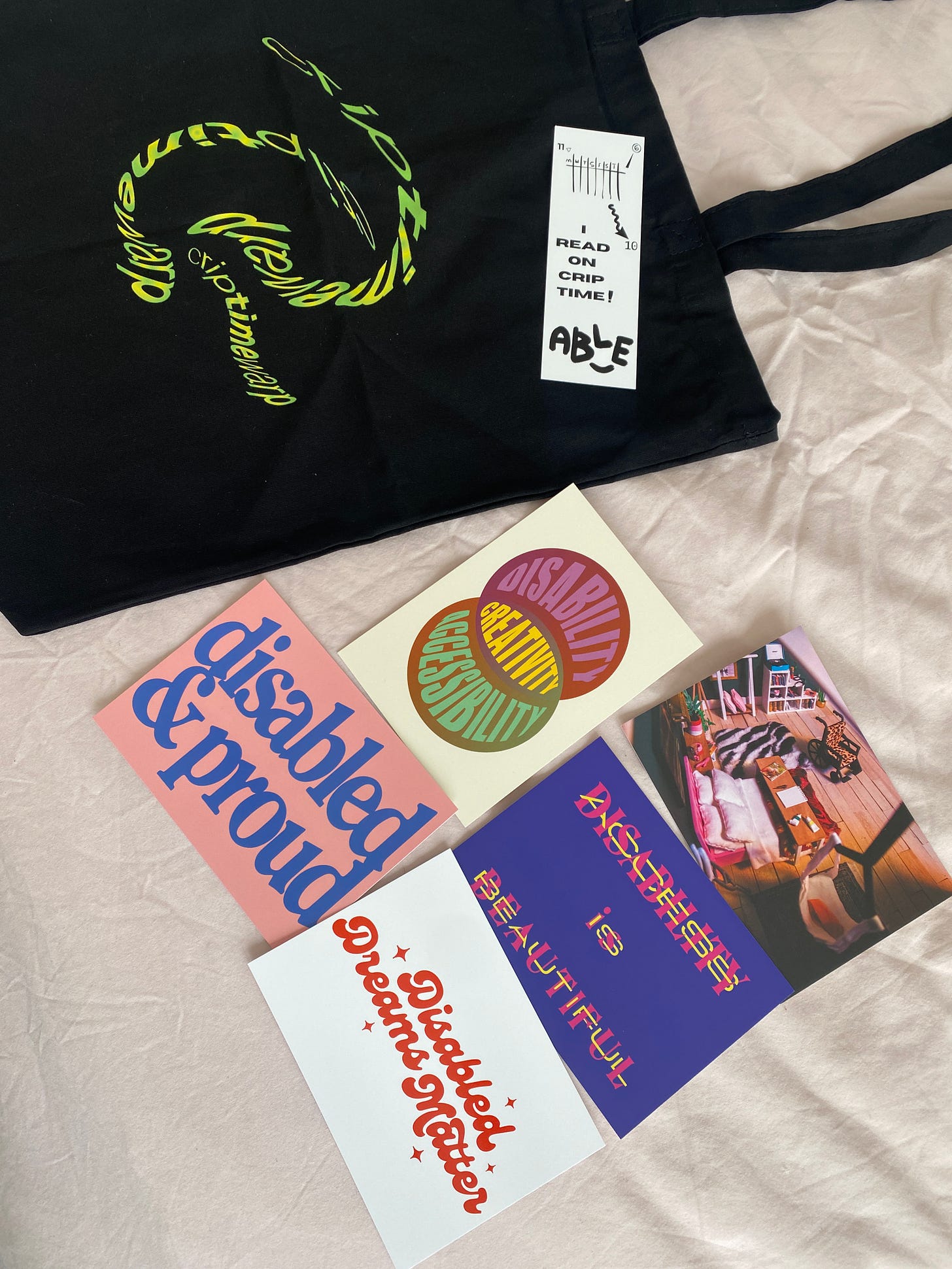A spread of postcards and merch on a pink bedspread. The postcards show graphic slogans such as “disabled and proud” and “disability/access is beautiful”. There is a bookmark resting on a black tote bag with a graphic that bends and curves reading “crip time warp”.