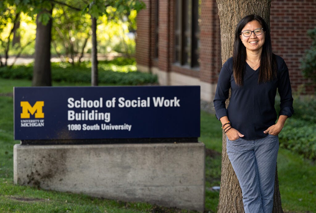 A photo of an Asian femme leaning against a tree near a sign for the School of Social Work Building at the University of Michigan.