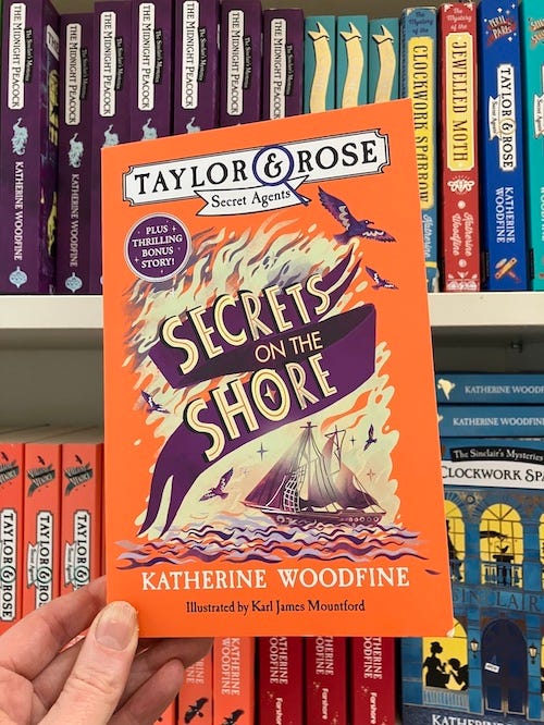 A copy of 'Taylor & Rose Secret Agents: Secrets on the Shore' with a bright orange cover, being held up in front of a bookshelf full of other books in the same series