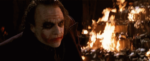the joker is standing in front of a fire and looking at it .