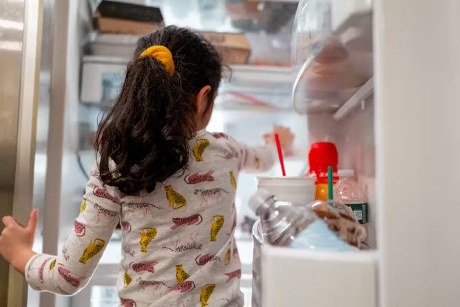 Little girl in pajamas reaches her hand into open refrigerator