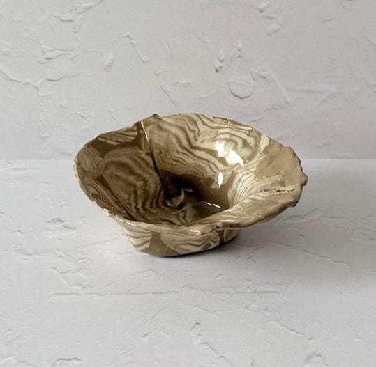 May include: A small ceramic bowl with a light brown and white marbled glaze. The bowl has a slightly irregular shape and a wide, shallow opening.