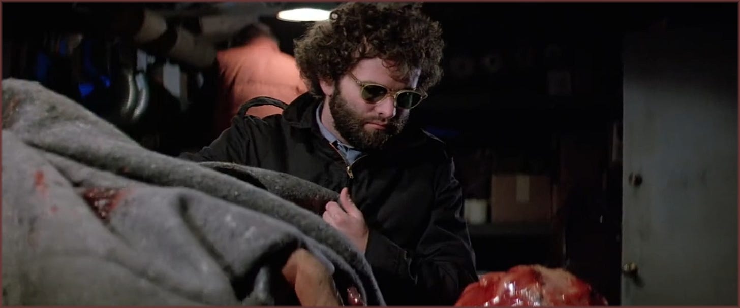 Film frame from The Thing (1982): Windows (Thomas G. Waites) leaning over the charred, split-faced remains of a creature recovered from the Norwegian camp, now laid out in the infirmary at Outpost 31. Expression a mixture of revulsion and fascination.