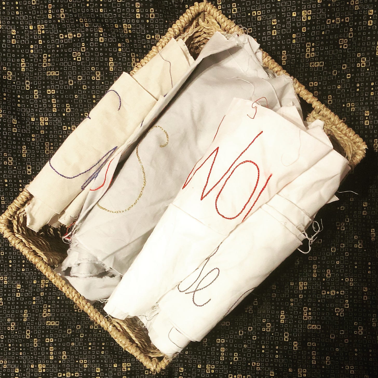 A basket of rolled fabric with some embroidered letters visible. The basket is placed on a piece of black and gold fabric